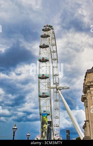Ein großes, weißes Riesenrad mit kreisförmigen Schoten steht vor einem dramatischen, bewölkten Himmel. Ein Teil eines Steingebäudes ist rechts in London zu sehen. Stockfoto