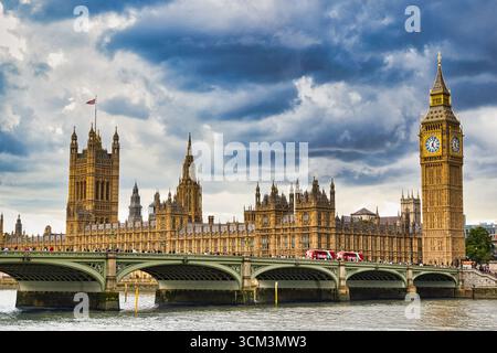Die Houses of Parliament und Big Ben stehen majestätisch vor einem dramatisch bewölkten Himmel. Eine Brücke überspannt einen Wasserkörper im Vordergrund, mit rotem Doub Stockfoto