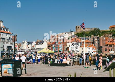 Eine geschäftige Uferszene mit einer Menschenmenge, Gebäuden und einer Union Jack Flagge vor einem klaren blauen Himmel in Whitby, Großbritannien. Stockfoto