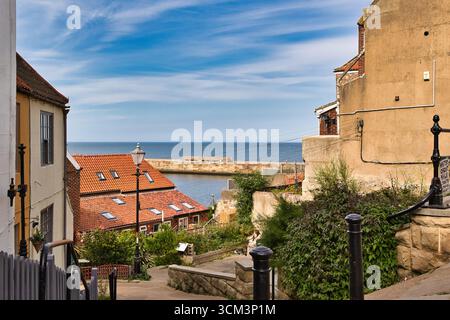 Ein steiler Pfad führt vorbei an Küstengebäuden in Richtung eines Hafens unter einem hellblauen Himmel mit verstreuten Wolken in Whitby, Großbritannien. Stockfoto