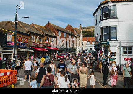 Eine geschäftige Straßenszene an einem sonnigen Tag, bei der Menschen an Geschäften entlang beiden Seiten der Straße unter einem hellblauen Himmel in Whitby, Großbritannien, vorbeilaufen. Stockfoto
