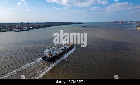 Ein großes Frachtschiff segelt auf einem breiten, trüben Gewässer und hinterlässt eine weiße Spur. Eine städtische Küste mit Gebäuden säumt auf der linken Seite, mit entferntem Por Stockfoto