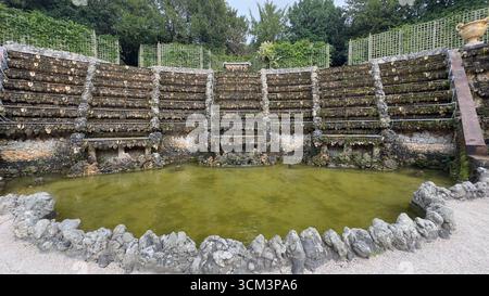 Ein kunstvoll verziertes, gestuftes Steingebäude mit einem Teich an der Basis, vor einer Kulisse aus üppigem Grün und Gitterzäunen. Stockfoto