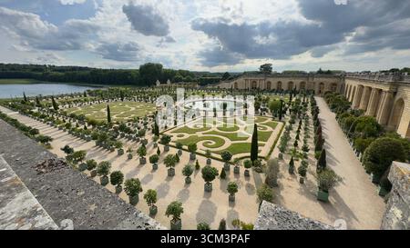 Ein großer Garten mit symmetrischen Parterres, einem runden Teich und einer Reihe von Topfbäumen, neben einem Steingebäude mit Bögen unter bewölktem Himmel. Stockfoto