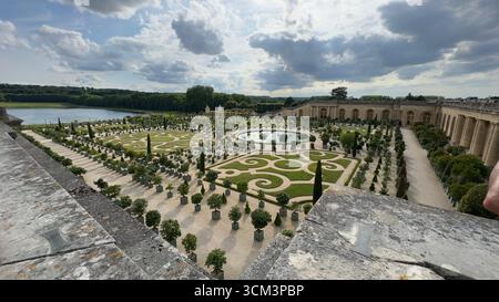 Ein Blick aus der Luft zeigt einen großen Garten mit gepflegten Rasenflächen, symmetrischen Mustern, einem Teich, Topfbäumen und einem langen, bogenförmiges Gebäude unter einer bewölkten Ske Stockfoto