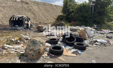 Ein ausgebranntes Auto liegt in einem mit Trümmern übersäten Außenbereich zwischen einem Haufen von Müll, Reifen und Felsen. Stockfoto