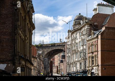 Ein Blick auf eine Straße, gesäumt von historischen Backsteingebäuden, mit einer gewölbten Eisenbahnbrücke in der Ferne unter einem teilweise bewölkten blauen Himmel in Newcas Stockfoto