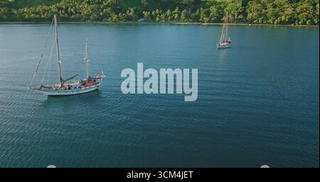 Fidschi-Inseln: Zwei Segelboote, die auf ruhigem blauem Wasser in der Nähe einer tropischen Küste schwimmen, üppige grüne Vegetation, die von einem warmen Sonnenuntergang beleuchtet wird. Wilde Naturreiselandschaft. Drohnenflug aus der Luft Stockfoto