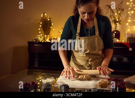 Eine Frau, die eine Schürze trägt, rollt Keksenteig in einer warmen Umgebung mit Weihnachtslichtern und festlichen Dekorationen im Hintergrund, um holi zu demonstrieren Stockfoto