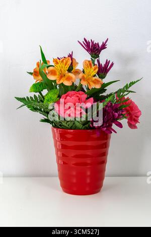 Strauß verschiedener Blumen in einer roten Vase auf einem weißen Tisch vor einer weißen Wand. Inneneinrichtung. Stockfoto