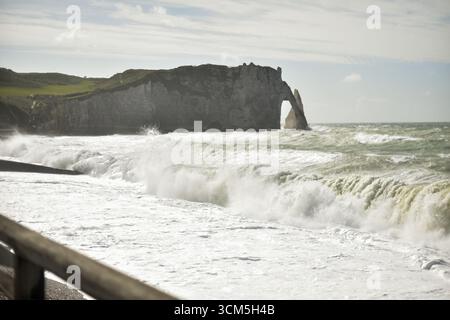 Dramatischer Blick auf die Klippe von Etretat unter strahlender Sonne und stürmischem Himmel. Mächtige, schaumige Wellen krachen gegen die Küste. Ein Holzzaun umrahmt die Szene und verleiht diesem wilden Moment an der Küste Tiefe. Stockfoto