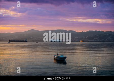 Farbenfroher Sonnenuntergang über der Bucht mit verankerten Frachtschiffen und Fischerbooten in Başiskele, Kocaeli, Türkiye Stockfoto