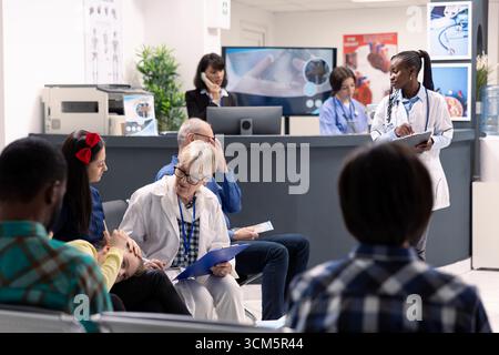 Beschäftigter Warteraum des Krankenhauses mit älterem männlichen Arzt, der Behandlungspläne mit Mutter und Tochter bespricht. Weibliche Pflegekräfte unterstützen Patienten und verwalten medizinische Unterlagen in der Nähe. Stockfoto