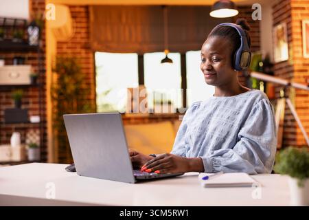 Schwarze Freelancerin genießt Musik über Kopfhörer, während sie auf einem digitalen Gerät schreibt. Glückliche afroamerikanische Frau mit schnurlosem Headset sitzt am Schreibtisch, studiert und bereitet eine Präsentation auf dem Laptop vor. Stockfoto
