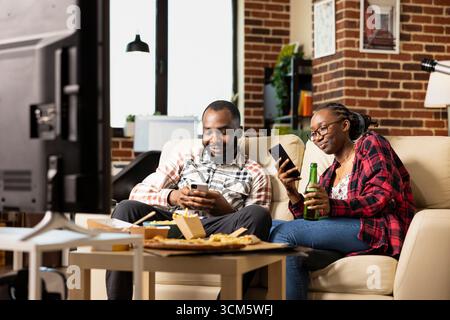 afroamerikanische Männer und Frauen saßen zusammen auf dem Sofa und konzentrierten sich auf ihre Smartphones. Modernes schwarzes Paar, das in sozialen Medien surft und in gemütlichem Wohnzimmer in Verbindung bleibt. Stockfoto