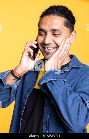 Indische Person lächelt während eines Gesprächs mit seiner Gruppe von Freunden, chelt auf einer Telefonleitung vor orangem Hintergrund. Junger Erwachsener mit einem Männerbrötchen in lässiger Kleidung, Klatsch. Stockfoto