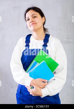 Junge Mechanikerin in blauen Overalls, die Arbeitsbücher hält Stockfoto