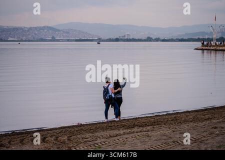 Zwei Freunde machen ein Selfie an der ruhigen Küste von Başiskele mit ruhigem Wasser und einer fernen Stadtlandschaft in der Abenddämmerung. Başiskele, Türkiye – 11. August 2023 Stockfoto