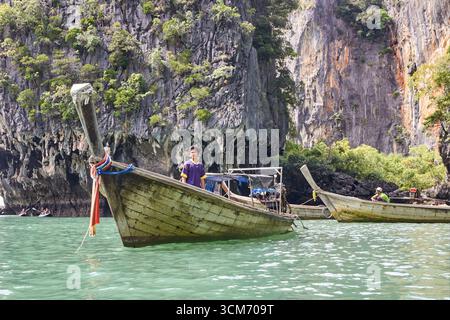 PHUKET, THAILAND - 12. NOVEMBER 2017: Asiatischer junger Mann auf einem traditionellen Holzboot inmitten von Kalksteinklippen, umgeben von türkisfarbenem Wasser. Stockfoto