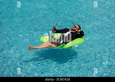 Geschäftsmann im Anzug mit Laptop im Swimmingpool. Verrückter Geschäftsmann schwimmt im Sommerurlaub. Aufgeregter Geschäftsmann in Neoprenanzug im Swimmingpool Stockfoto