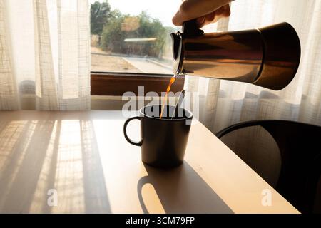 Kaffee wird aus einer Mokakanne in eine Tasse gegossen, morgens kommt Sonnenlicht durch das Fenster Stockfoto