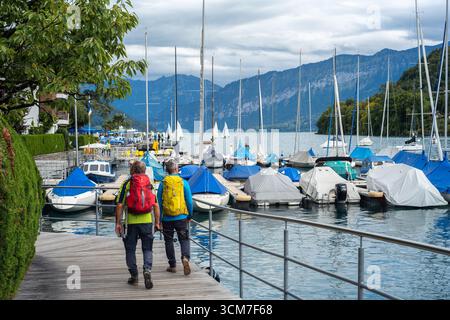 Spiez, Schweiz - 11. September 2025: Zwei Wanderer, die an einem Jachthafen in Spiez mit Booten und Bergen im Hintergrund spazieren. Stockfoto