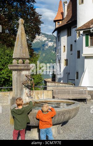 Spiez, Schweiz - 11. September 2025: Zwei Kinder spielen in der Nähe eines Steinbrunnens von Schloss Spiez mit malerischer Bergkulisse. Stockfoto
