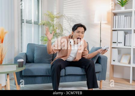 Ein junger Mann sitzt auf einem Sofa mit einem Game-Controller, voll konzentriert und frustriert, während er ein Videospiel spielt. Seine intensive Reaktion ist hochgradig Stockfoto