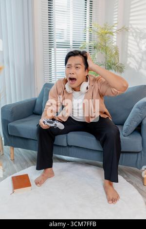 Ein junger Mann sitzt auf einem Sofa mit einem Game-Controller, voll konzentriert und frustriert, während er ein Videospiel spielt. Seine intensive Reaktion ist hochgradig Stockfoto