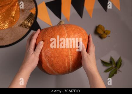 Eine Person hält einen orangefarbenen Kürbis in beiden Händen. Ein Hexenhut, ein schwarz-oranges Wimpelbanner und Herbstblätter stehen im Hintergrund auf der grauen Oberfläche. Halloween-Vorbereitungen sind im Gange. Stockfoto