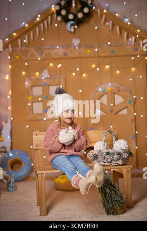 Entzückendes blondes Kind in rosa Strickpullover und weißer Beanie, das auf einer Holzbank neben einem verzierten Weihnachtsbaum mit Feenlichtern sitzt Stockfoto