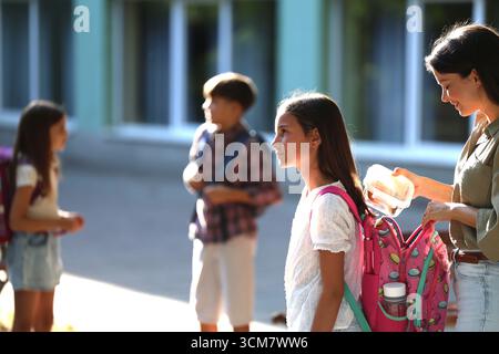 Mutter legt ihre Tochter in den Rucksack. Sie genießen die gemeinsame Zeit auf dem Schulhof während der Nachmittagsstunden Stockfoto
