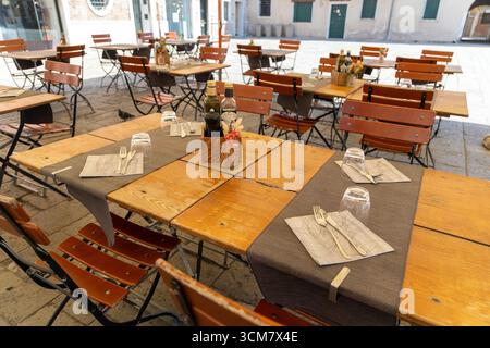 Leere Restauranttische im Freien auf dem sonnigen Platz. Ordentlich gedeckte Holztische und -Stühle in einem Restaurant im Freien auf einem ruhigen europäischen Platz, bereit für Stockfoto