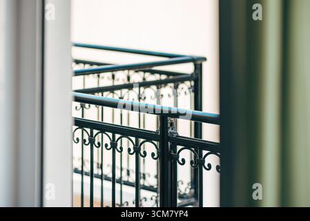 Elegantes, schwarzes schmiedeeisernes Balkongeländer, das durch ein offenes Fenster mit weichem Fokus betrachtet wird, bietet architektonische Details und ein Gefühl für eine ruhige Stadt Stockfoto