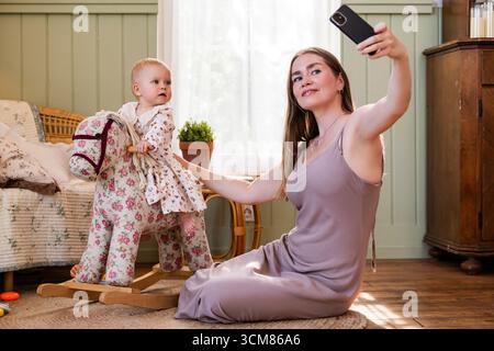 Mom macht ein Selfie mit ihrem Baby, das auf einem Schaukelpferd sitzt Stockfoto