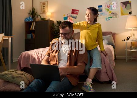 Kaukasischer Mann mittleren Alters sitzt auf dem Boden mit Laptop, während das kaukasische Mädchen dahinter steht und die Papierkrone auf dem Kopf in einem gemütlichen, mit Zeichnungen verzierten Schlafzimmer platziert Stockfoto