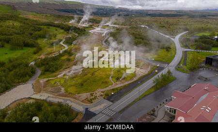 Luftbild von Haukadalur in Island mit dampfenden Lüftungsöffnungen, sprudelnden heißen Quellen, gewundenen Wegen, üppigem Grün und einem rot überdachten Gebäude. Stockfoto