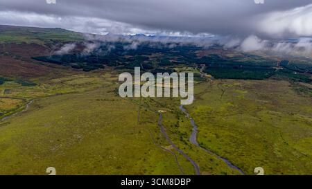 Panoramablick auf das grüne moosige Gelände Islands aus der Vogelperspektive mit gewundenen Flüssen, verstreuten Wäldern, fernen Bergen und einer kleinen Straße im Vorland Stockfoto