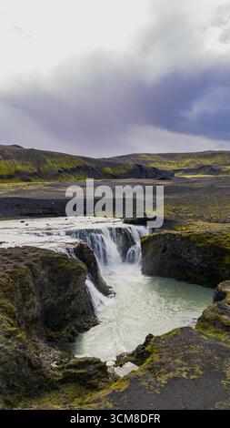 Ein kleiner Wasserfall fließt in einen türkisfarbenen Pool, umgeben von vulkanischen Klippen und grünem Moos. Die Luftaufnahme fängt das zerklüftete isländische Gelände ein. Stockfoto