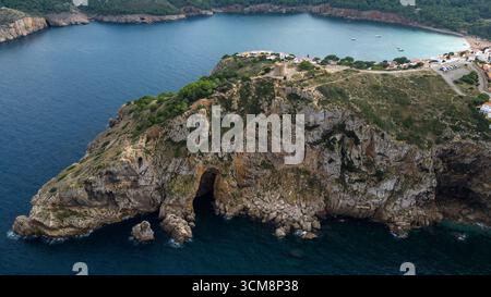 Das Bild zeigt die markante Kalksteinklippe von Montgo in der Nähe von L'Escala an der Costa Brava mit dem historischen Wachturm Torre de Montgo auf dem Hügel. Im Vordergrund ragen felsige Klippen und eine Meereshöhle spektakulär in das tiefblaue Mittelmeer, während im Hintergrund eine geschützte Bucht mit Booten und weißen Häusern zu sehen ist. Die Szene verbindet wilde Natur, historische Architektur und eine mediterrane Küstenlandschaft. Stockfoto