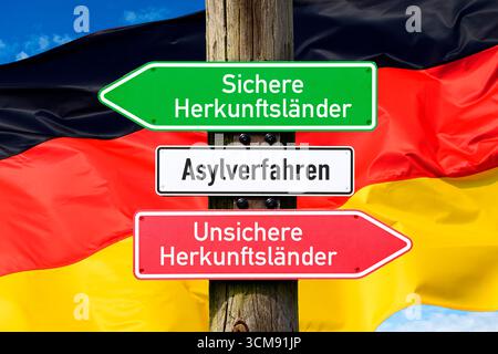 Wegweiser mit Aufschriften Asylverfahren, sichere Herkunftsländer und unsichere Herkunftsländer vor der deutschen Flagge, Fotomontage Stockfoto