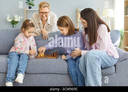 Kinder spielen Schach auf dem Sofa, während sie lächelnd von Mutter und Großmutter beobachtet und unterstützt werden. Stockfoto