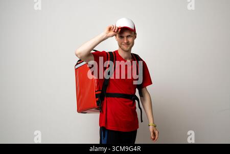 Lächelnder junger Liefermann in roter Uniform mit thermischem Rucksack auf weißem Atelierhintergrund Stockfoto