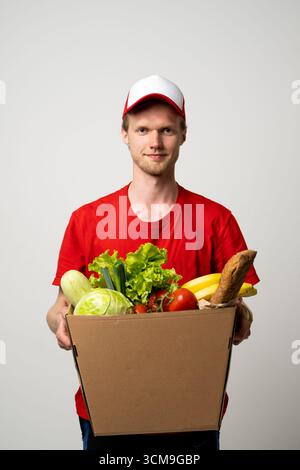 Junger Liefermann in roter Uniform, der Pappkarton voll mit frischem Gemüse und Obst hält Stockfoto