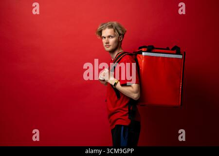 Junger Kuriermann in rotem T-Shirt mit Thermorucksack, der drinnen auf rotem Hintergrund steht Stockfoto