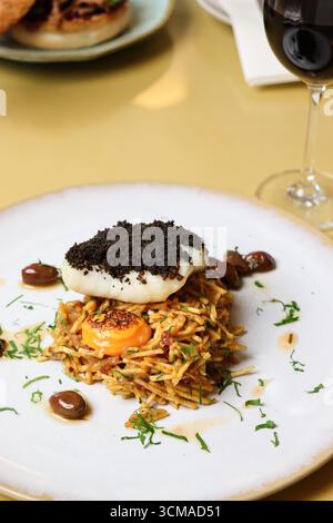 Gourmet-Gericht mit gebratenem Weißfisch, belegt mit schwarzen Krümeln, serviert über knusprigen Pommes mit einem reichhaltigen Eigelb und garniert mit Kräutern Stockfoto