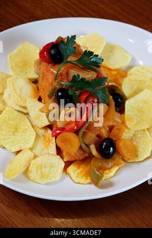 Traditioneller Kabeljau, das Hauptfischstück ist bedeckt mit einer bunten Mischung aus gebratenen Zwiebeln und Paprika, Kartoffelchips, garniert mit ganzer schwarzer Olive Stockfoto