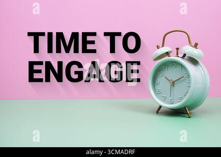 Schwarzer Wecker auf pastellgrünem und rosafarbenem Hintergrund mit dem Text „Time to Engage“ Stockfoto
