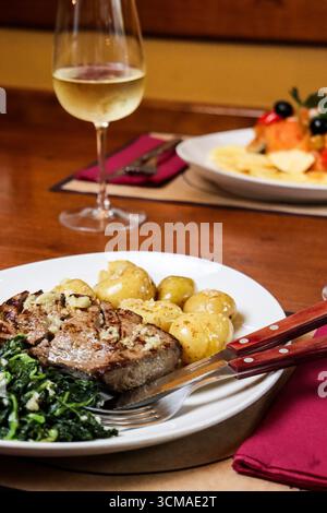 Nahaufnahme eines gegrillten Steaks mit sautiertem Spinat und gekochten Kartoffeln auf einem weißen Teller. Traditionelle herzhafte Mahlzeit begleitet von einem Glas Stockfoto