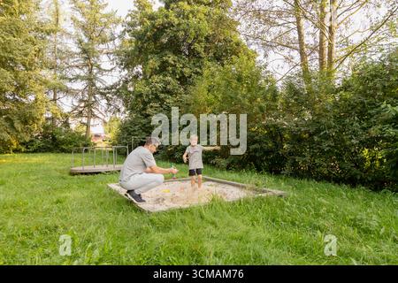 Ein Vater und sein Kind haben Spaß auf einem Spielplatz und nehmen an Aktivitäten in einem Sandkasten teil, umgeben von üppigem Grün. Ihr Lachen erfüllt die Luft Stockfoto